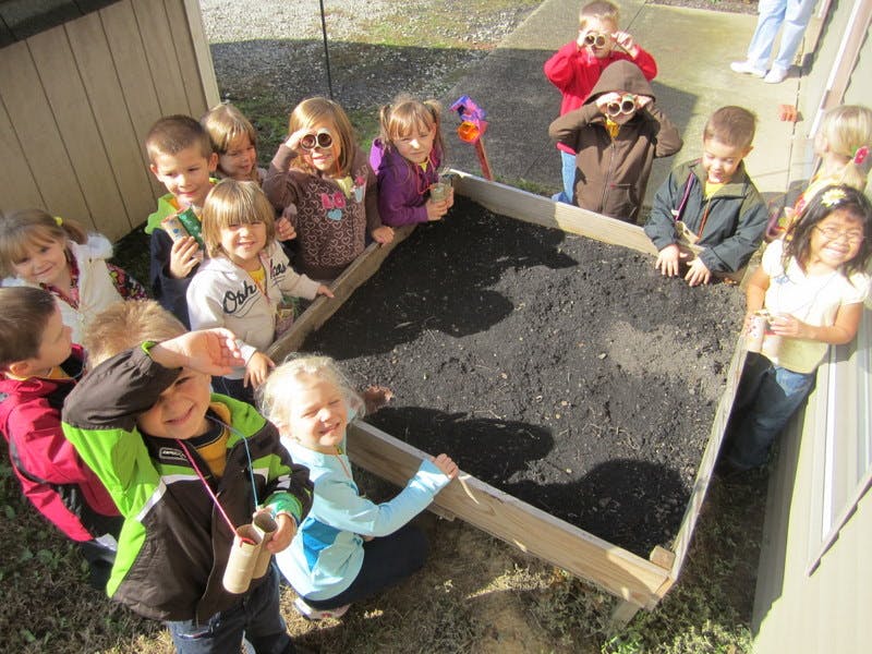 Children gathered around a raised garden bed filled with soil