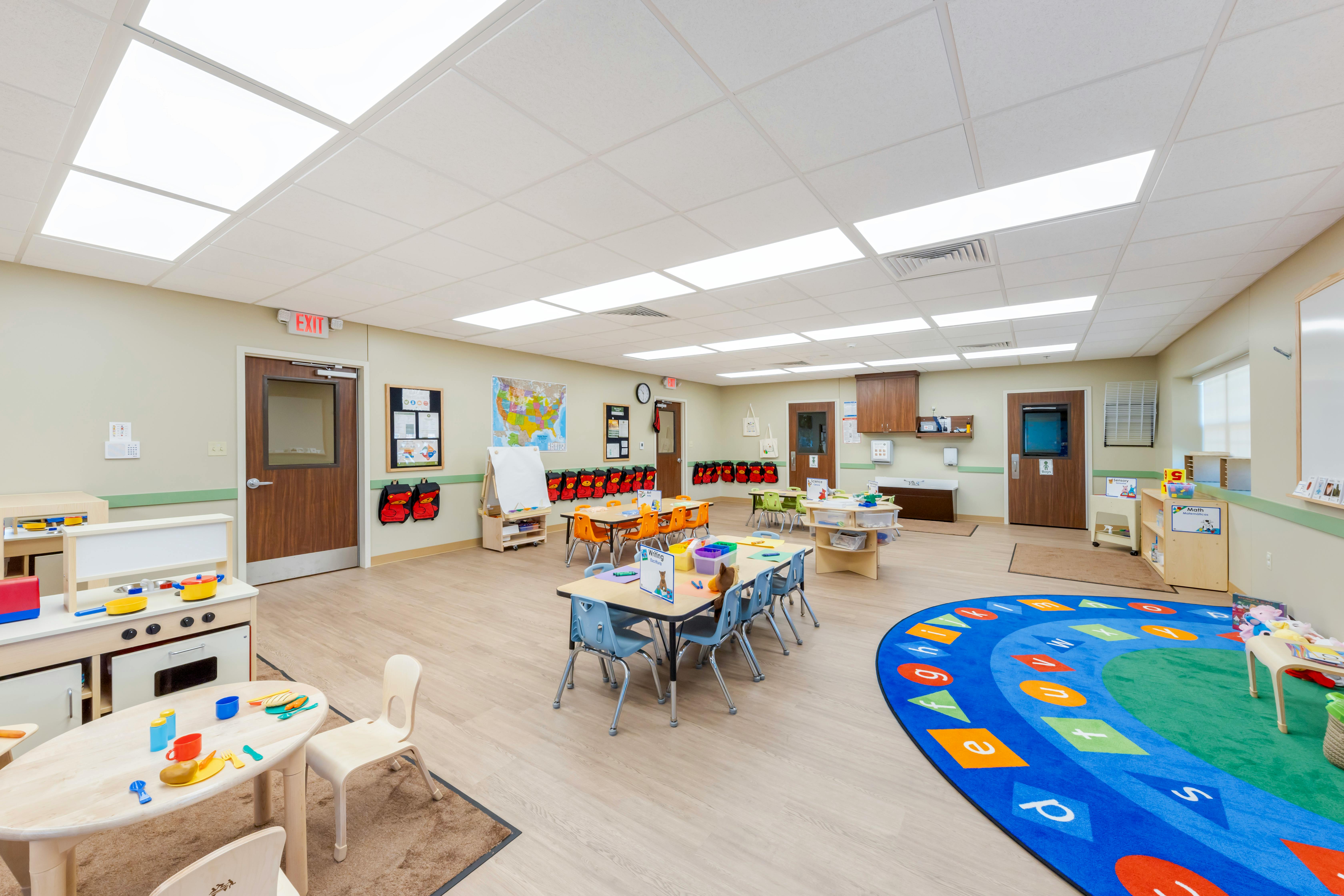 Empty classroom with small tables and chairs, play kitchen, and alphabet rug