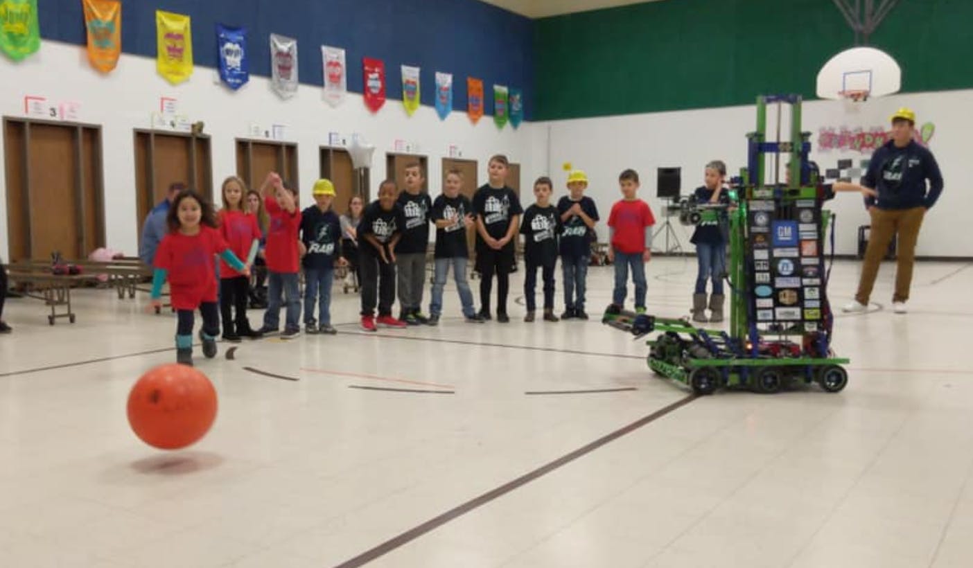Child kicking a ball in a gym with other children and a robot watching