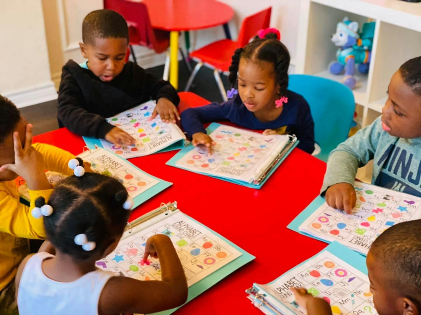 Children sitting at a red table, looking at papers with colorful shapes