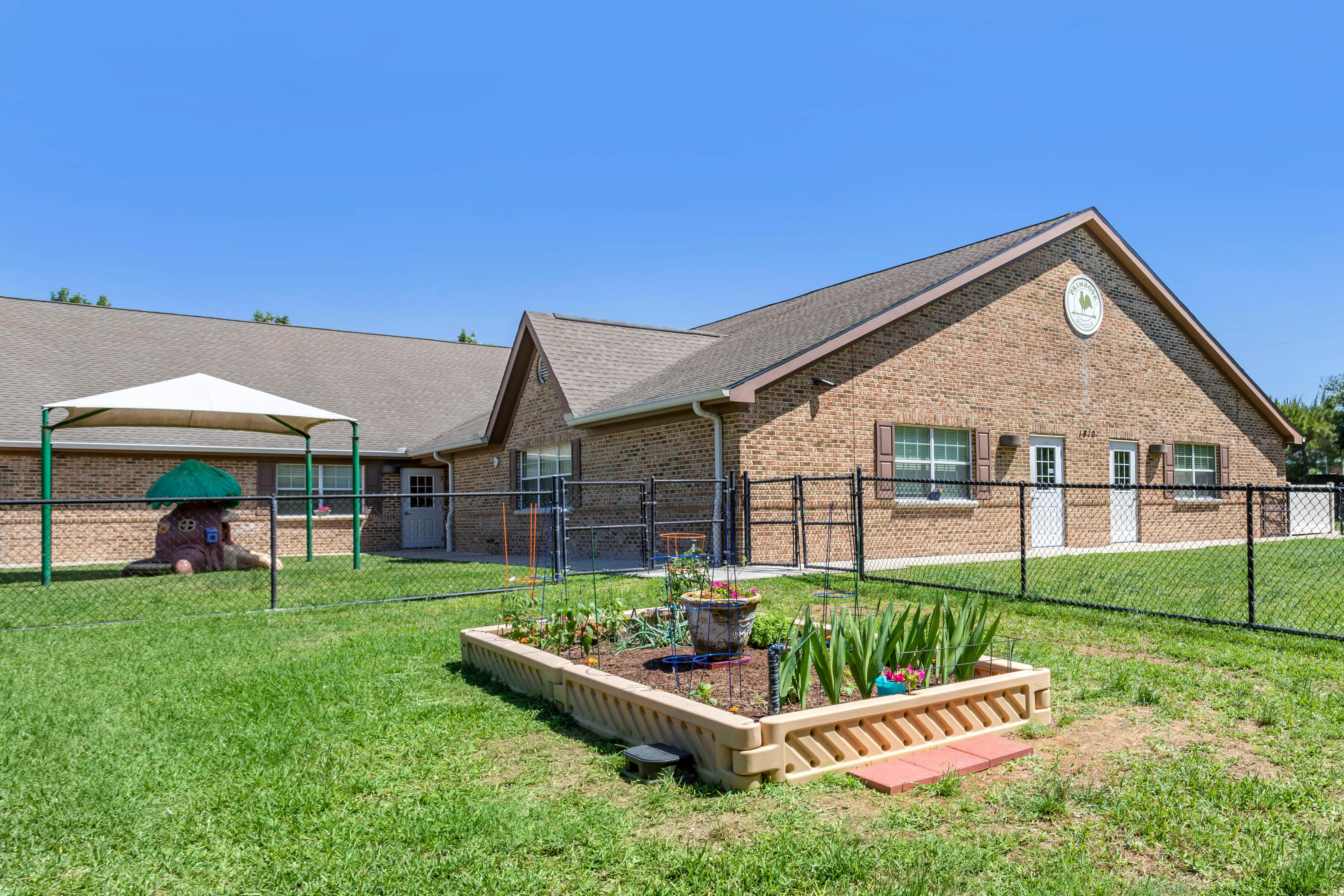 Fenced playground with play structure and garden bed in front of a brick building