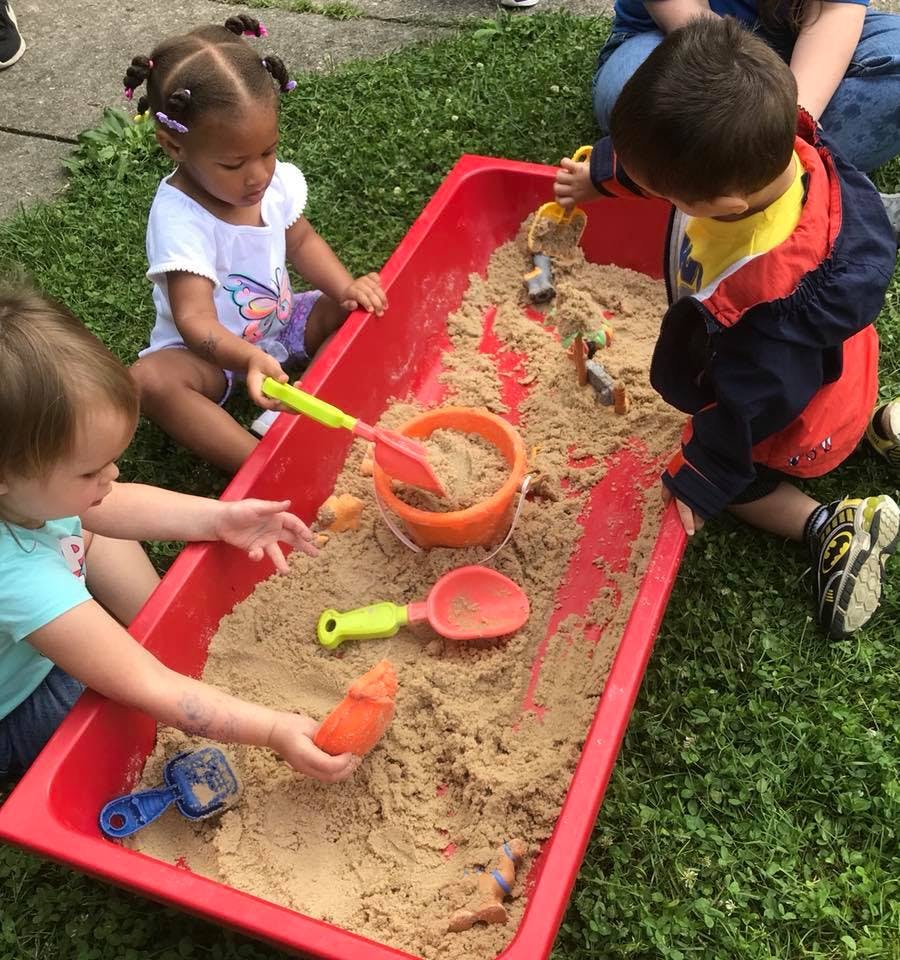 Three children playing in a red sandbox with sand toys