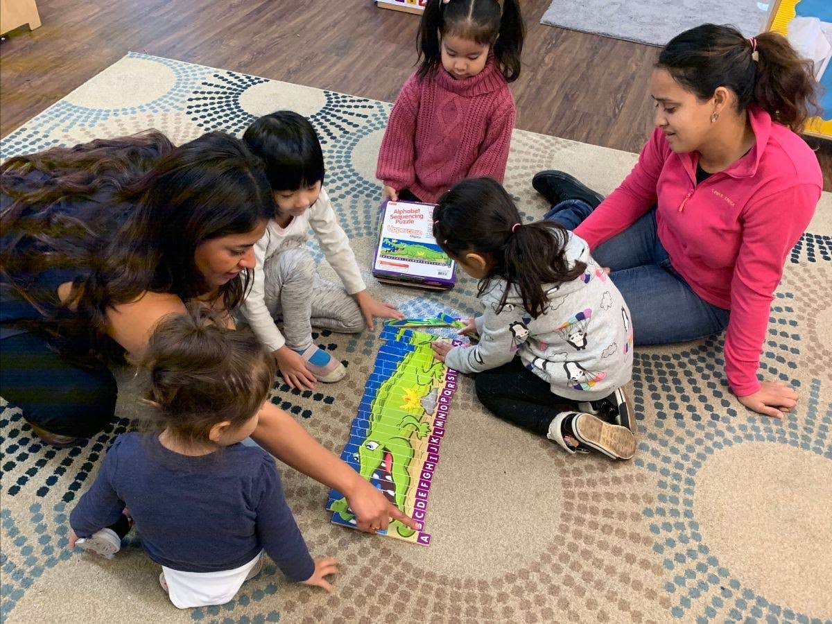 Two adults and four children playing with a large green alligator puzzle on the floor