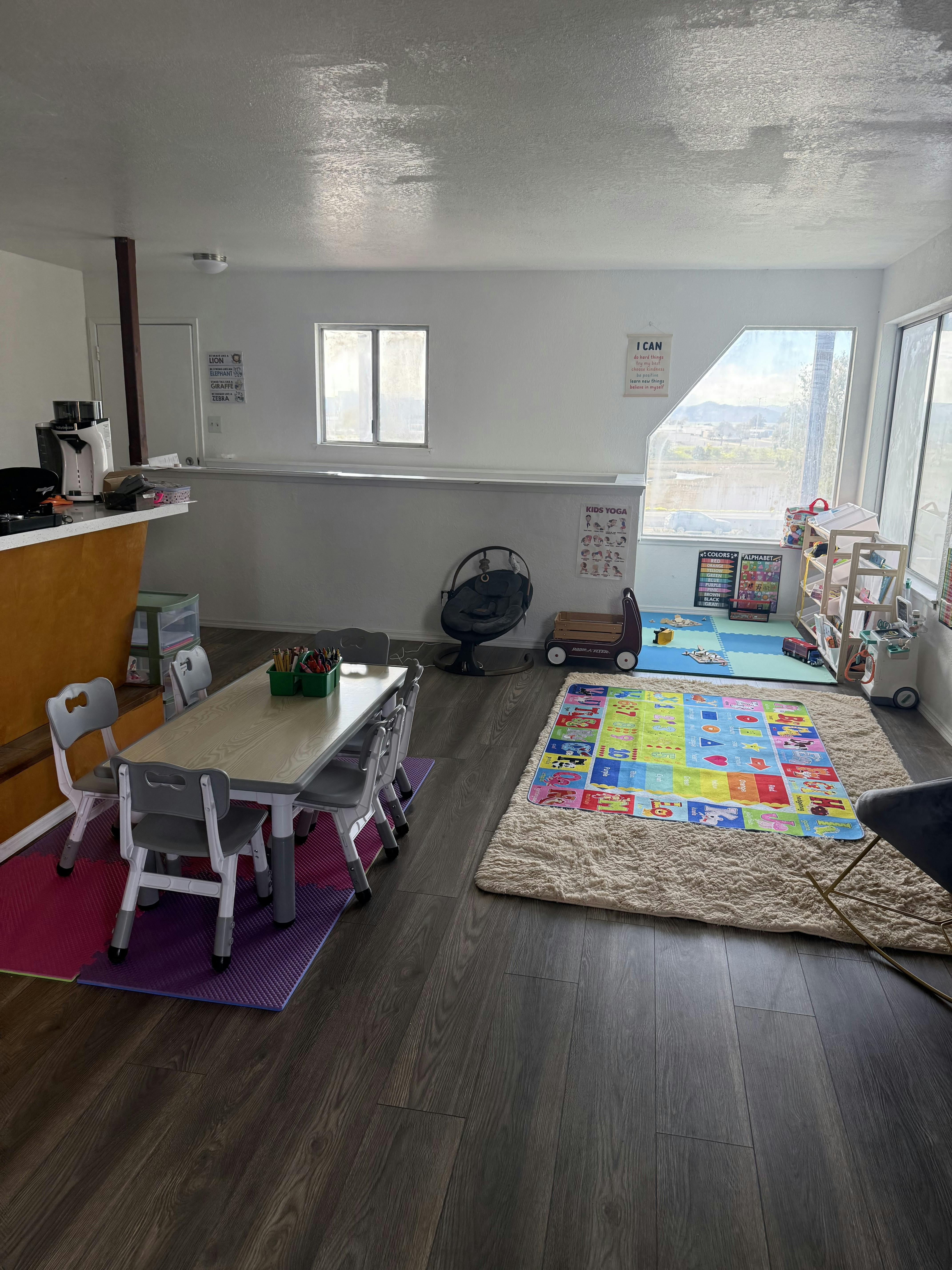 A room with a small table and chairs, a play mat, and large windows