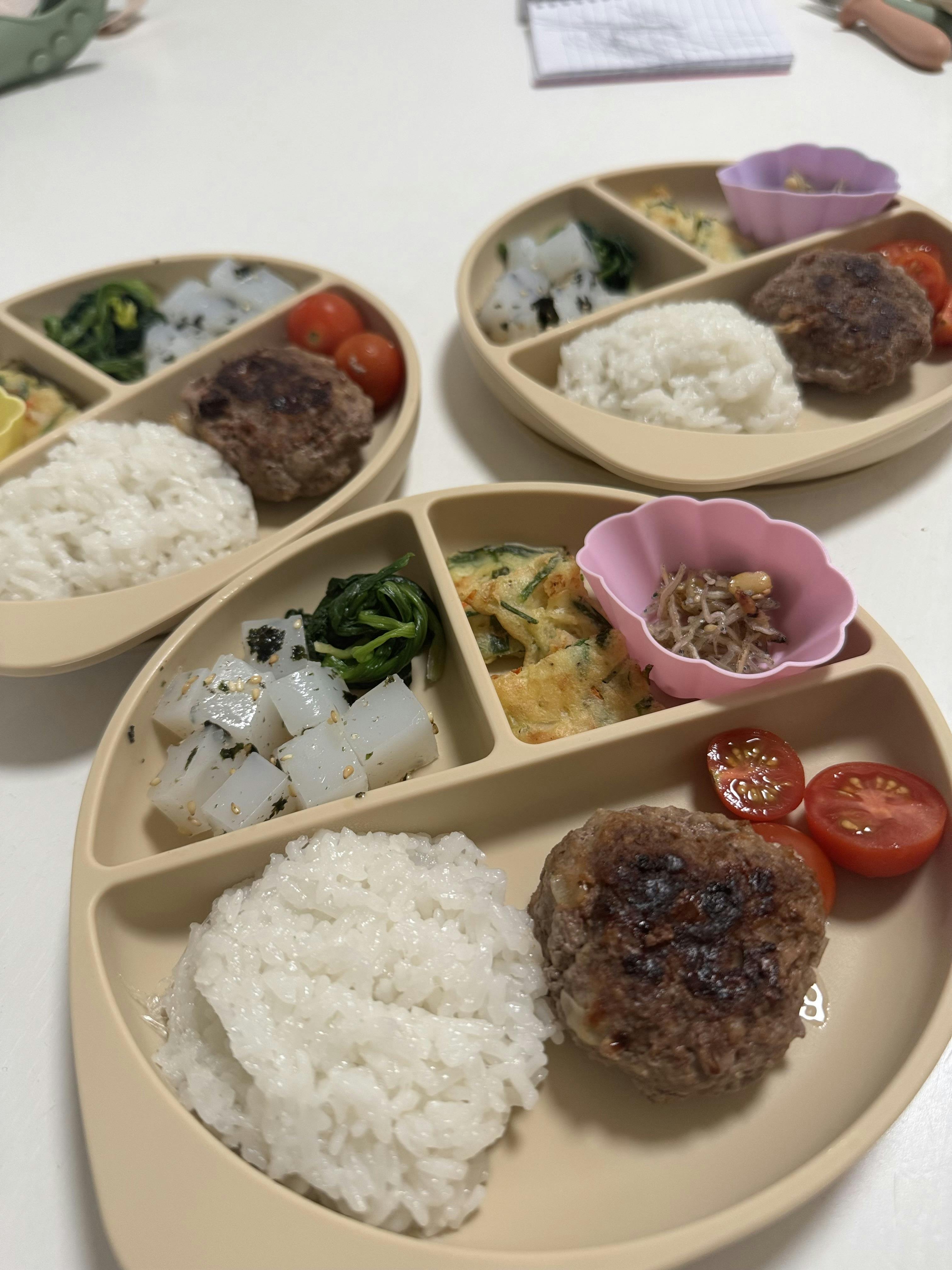 Three partitioned plates with rice, hamburger patties, cherry tomatoes, and other dishes