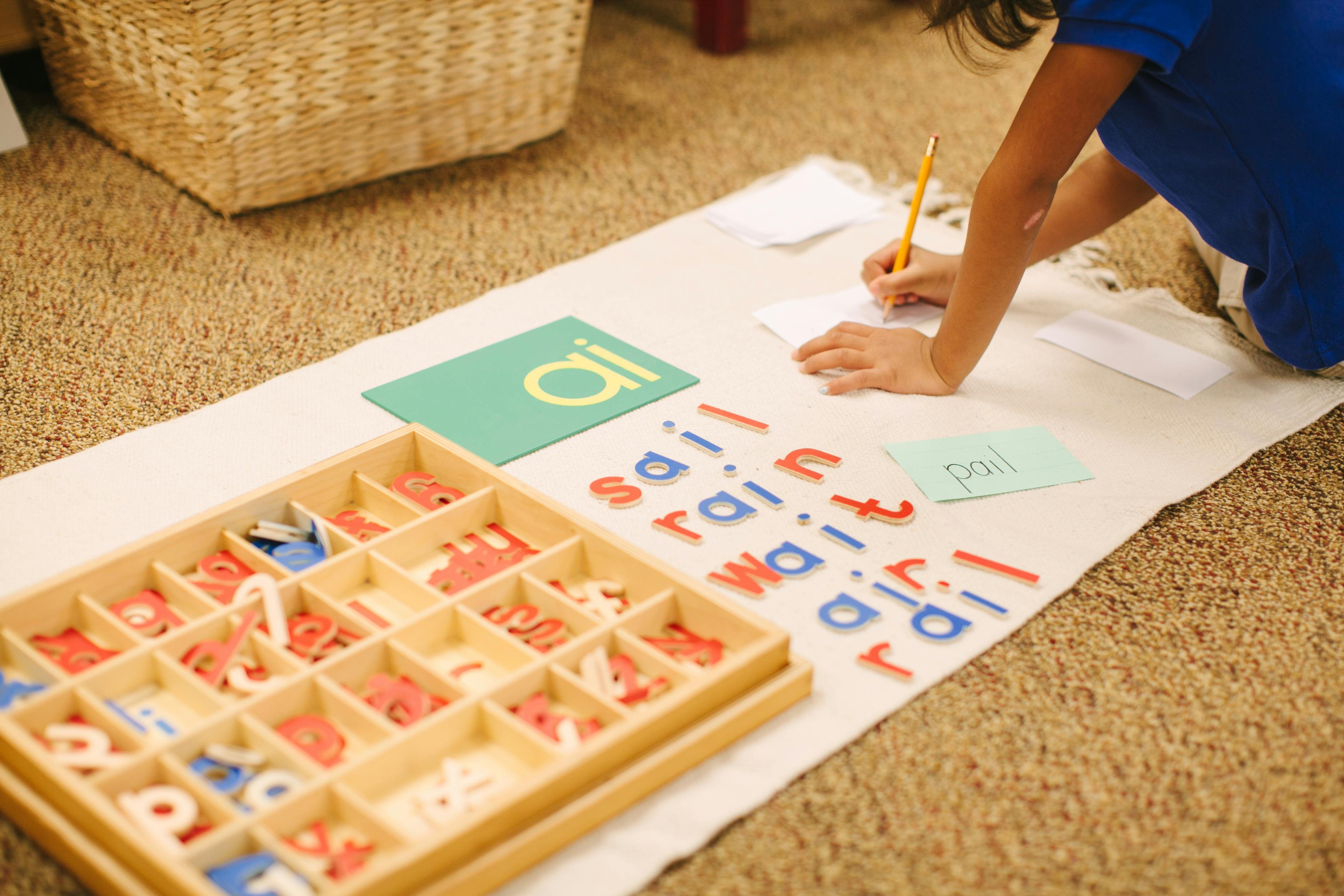 Child writing on paper with letter tiles and a letter box nearby