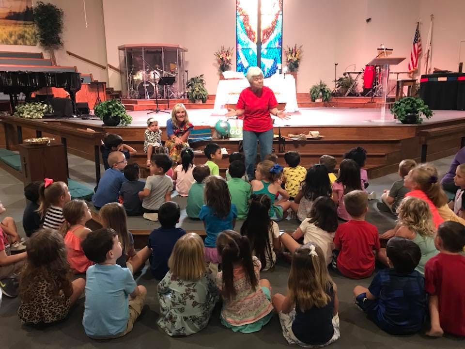 Adults speaking to a group of children seated on the floor in a large room