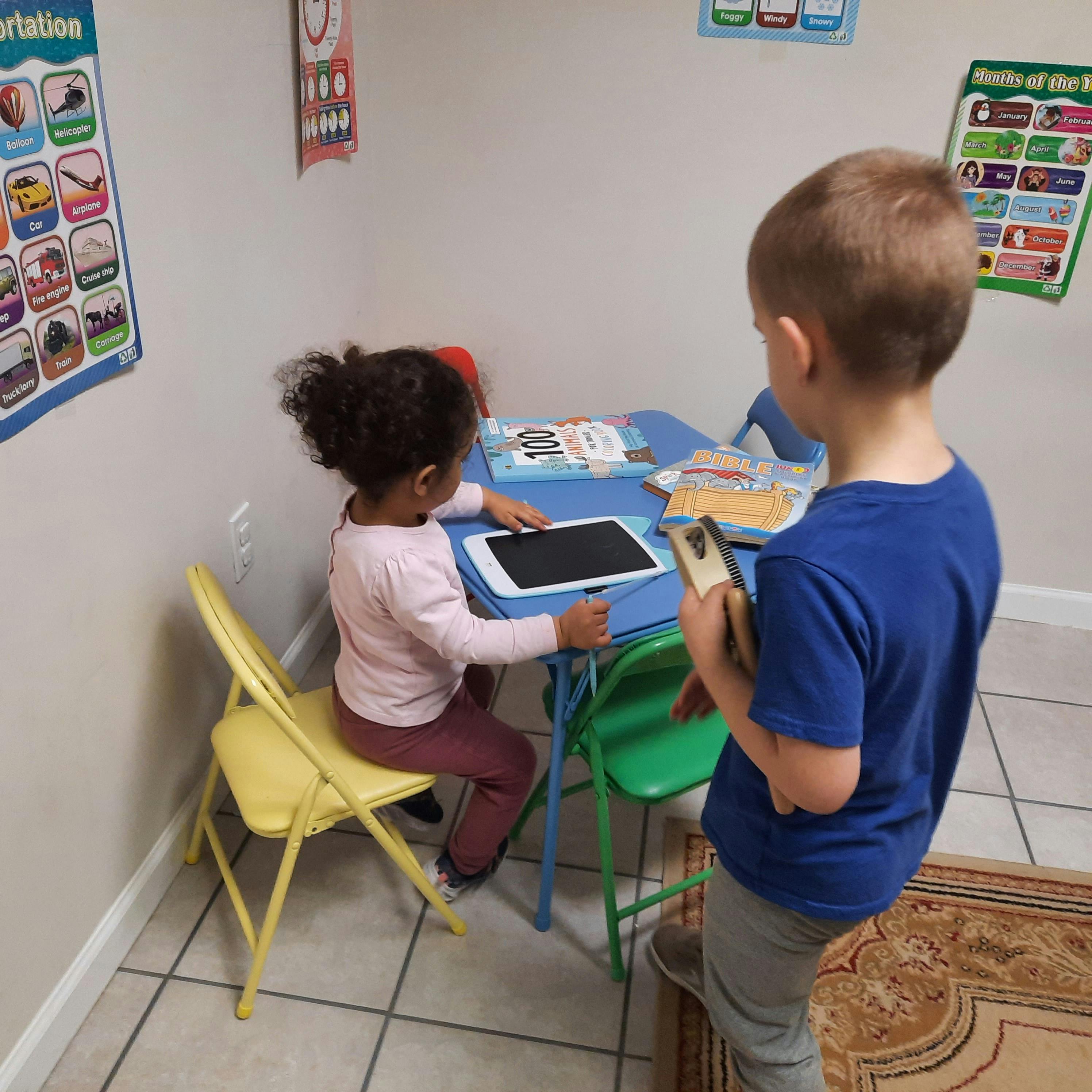 Child at table using a tablet, another child standing nearby