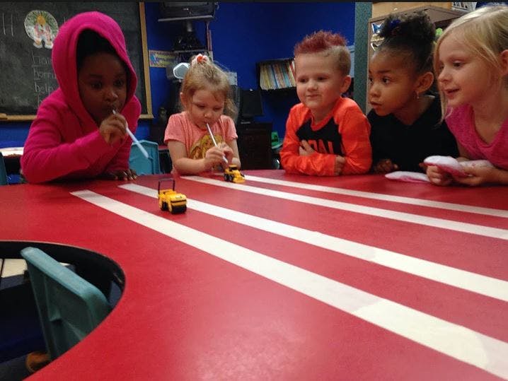 Children blowing through straws at toy cars on a red and white striped table