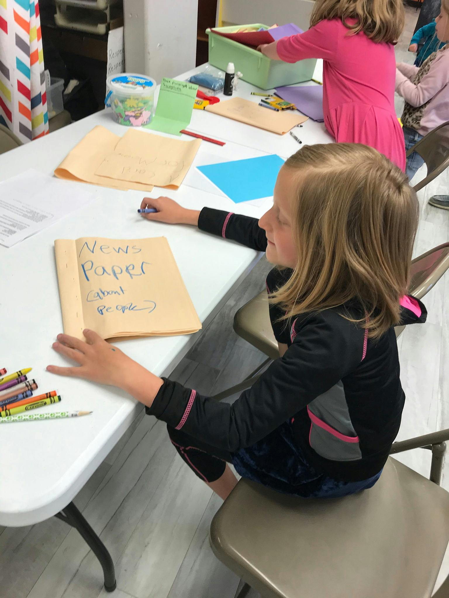 Child sitting at a table with a folder labeled "News Paper"
