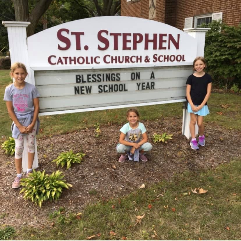 Three children posing in front of a sign for St. Stephen Catholic Church & School