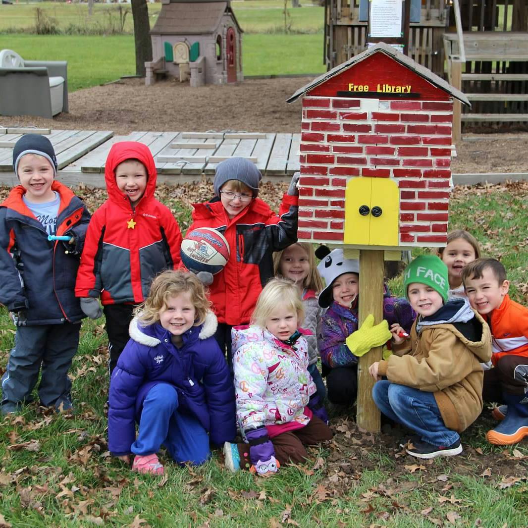 Group of children gathered around a small red brick structure with "Free Library" text