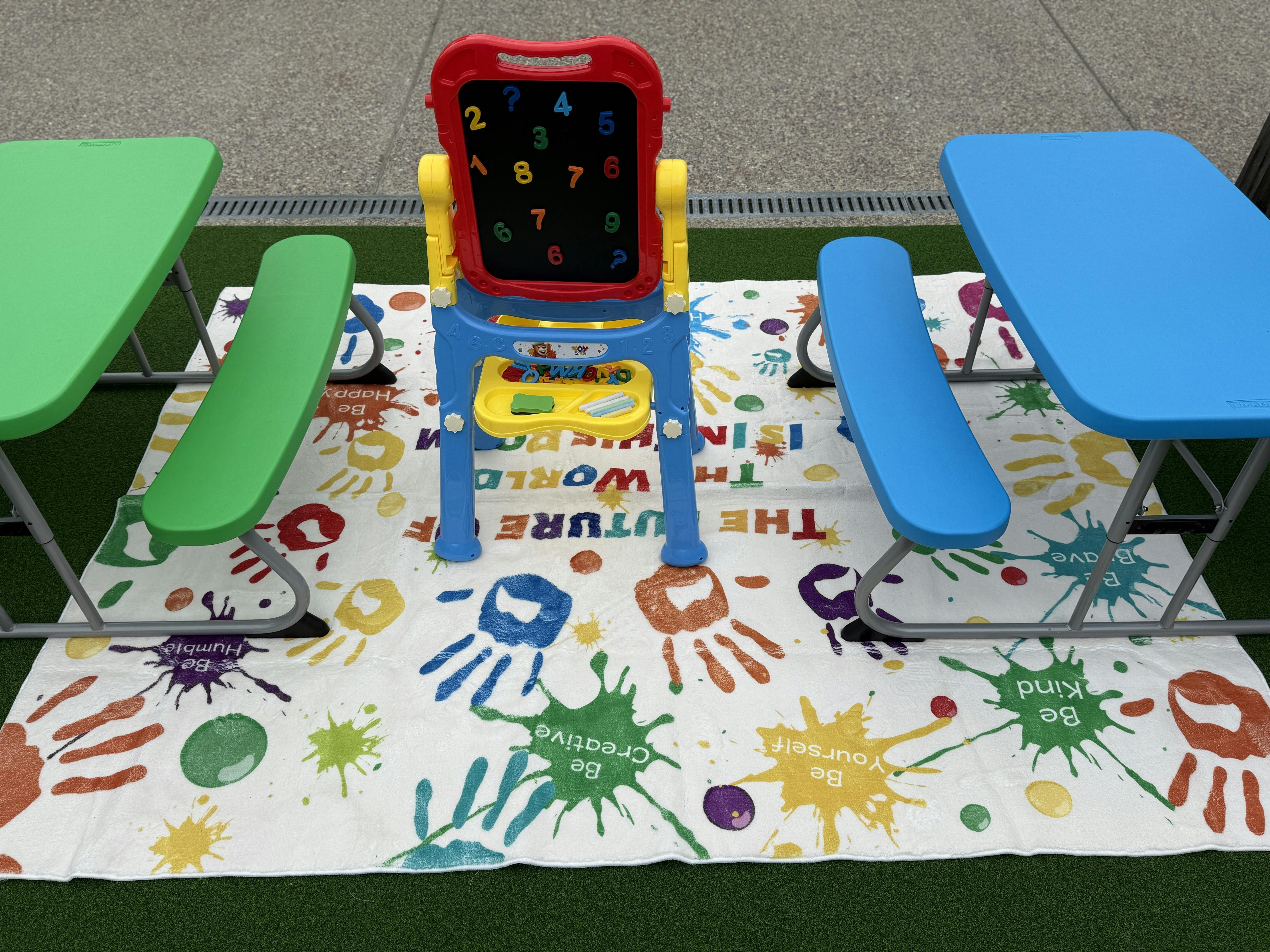 Children's play area with colorful tables, benches, and an easel on a mat