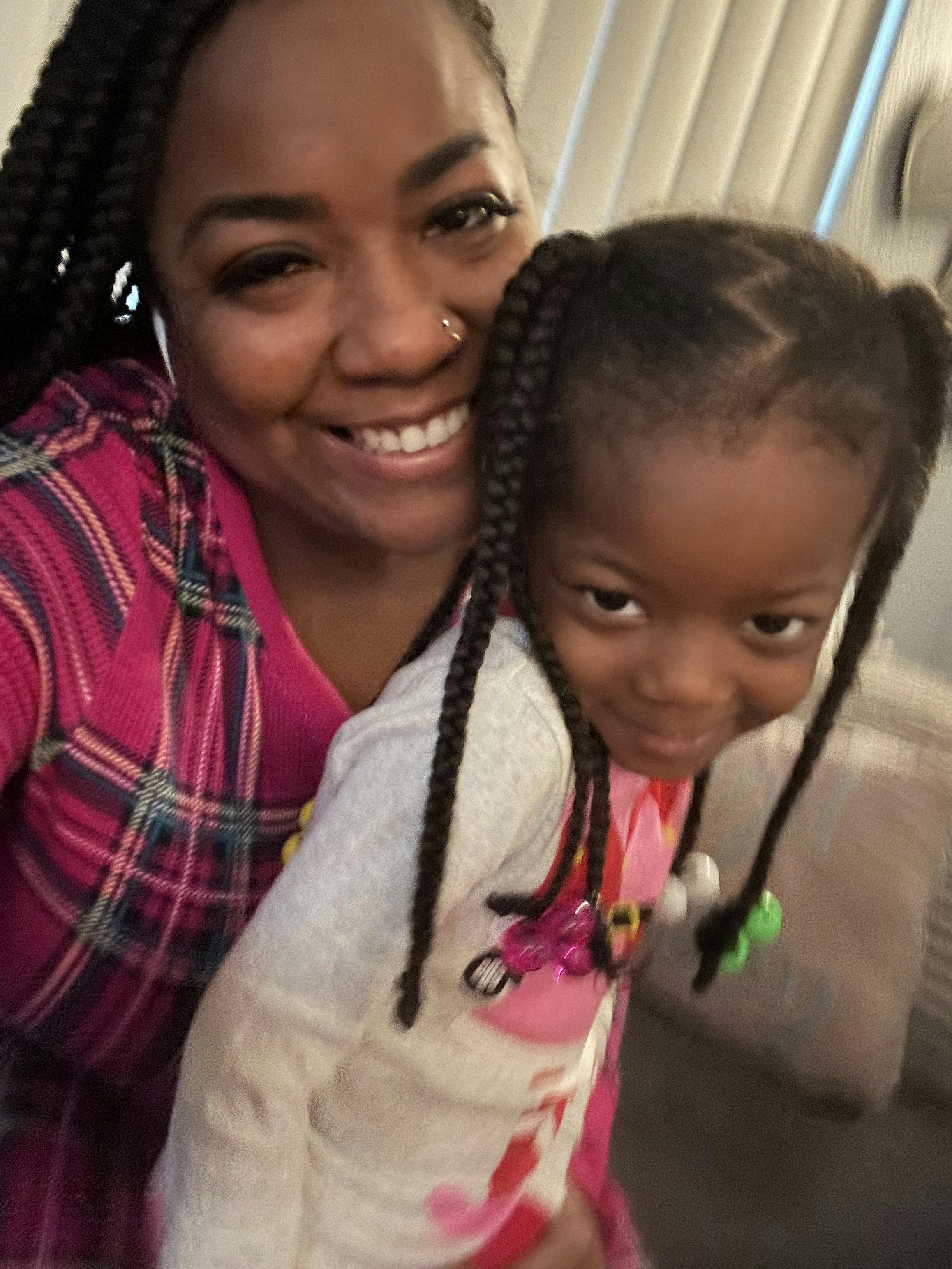 Smiling person wearing a plaid shirt holding a child with braided hair