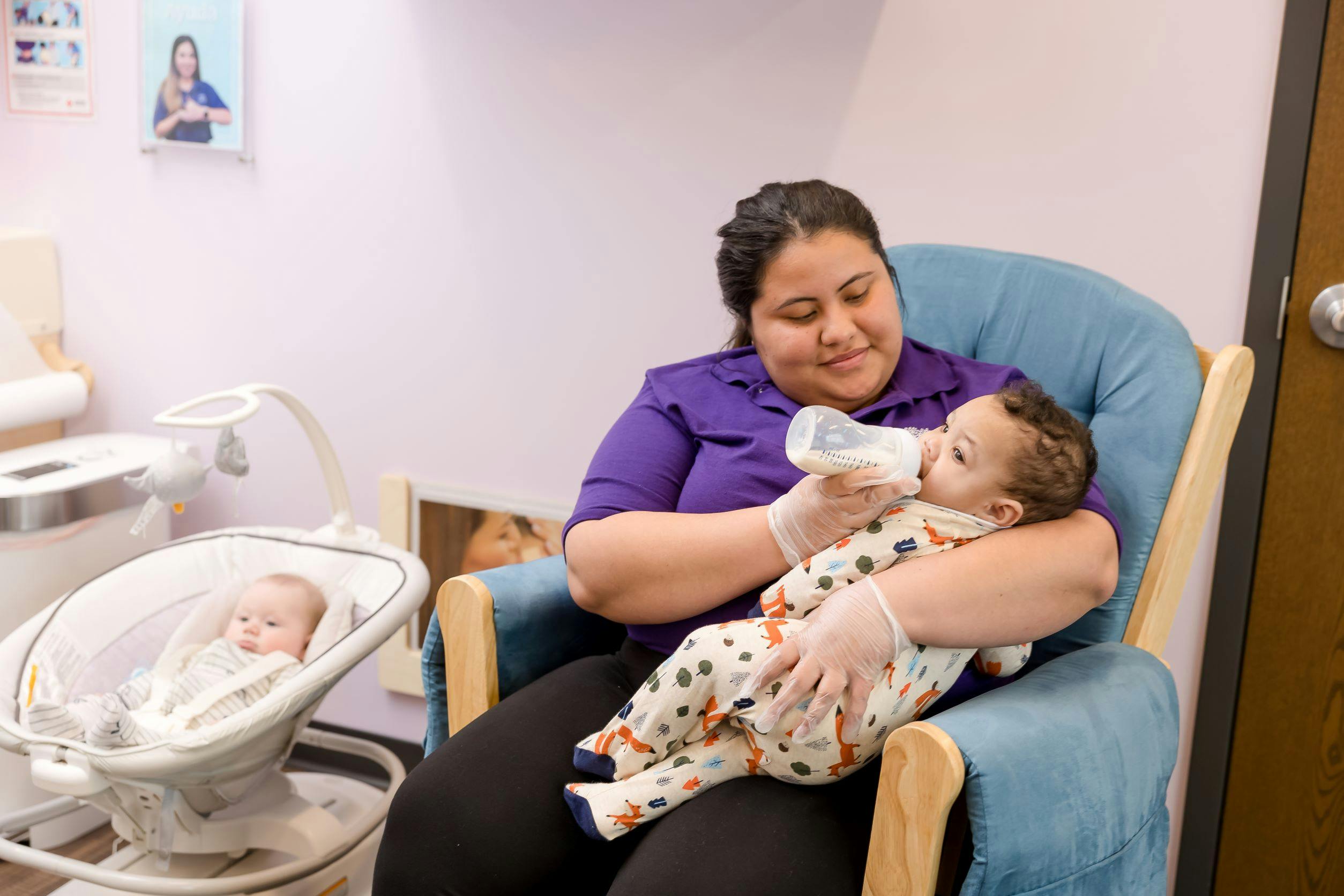 Person in gloves bottle-feeding a baby in a chair