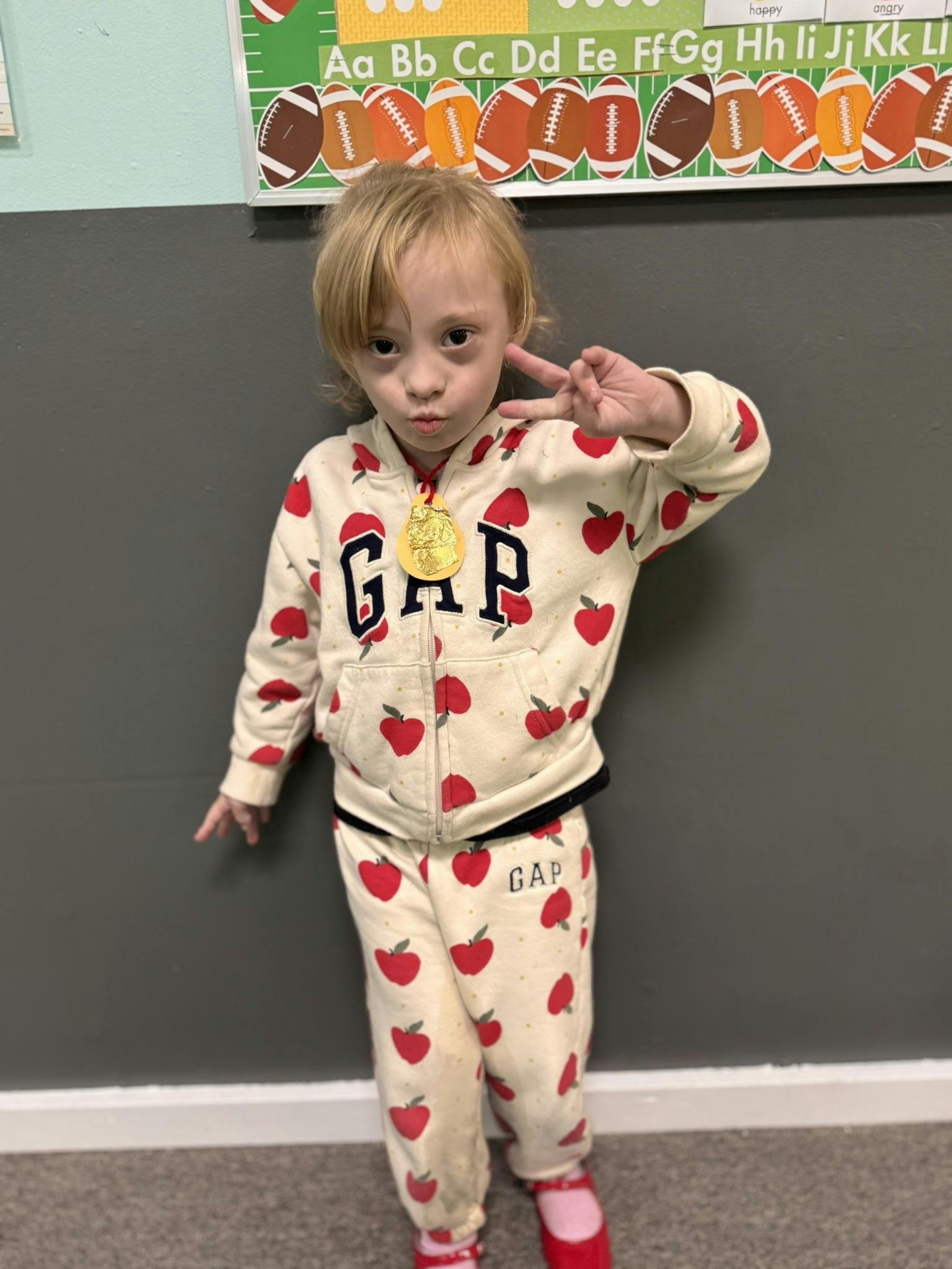 Child wearing an apple print outfit and a gold medal making a peace sign