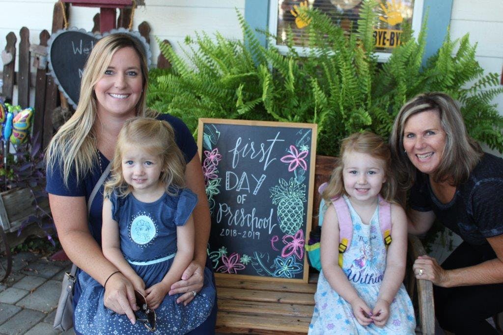Two people with two children and a chalkboard sign that says "First Day of Preschool"