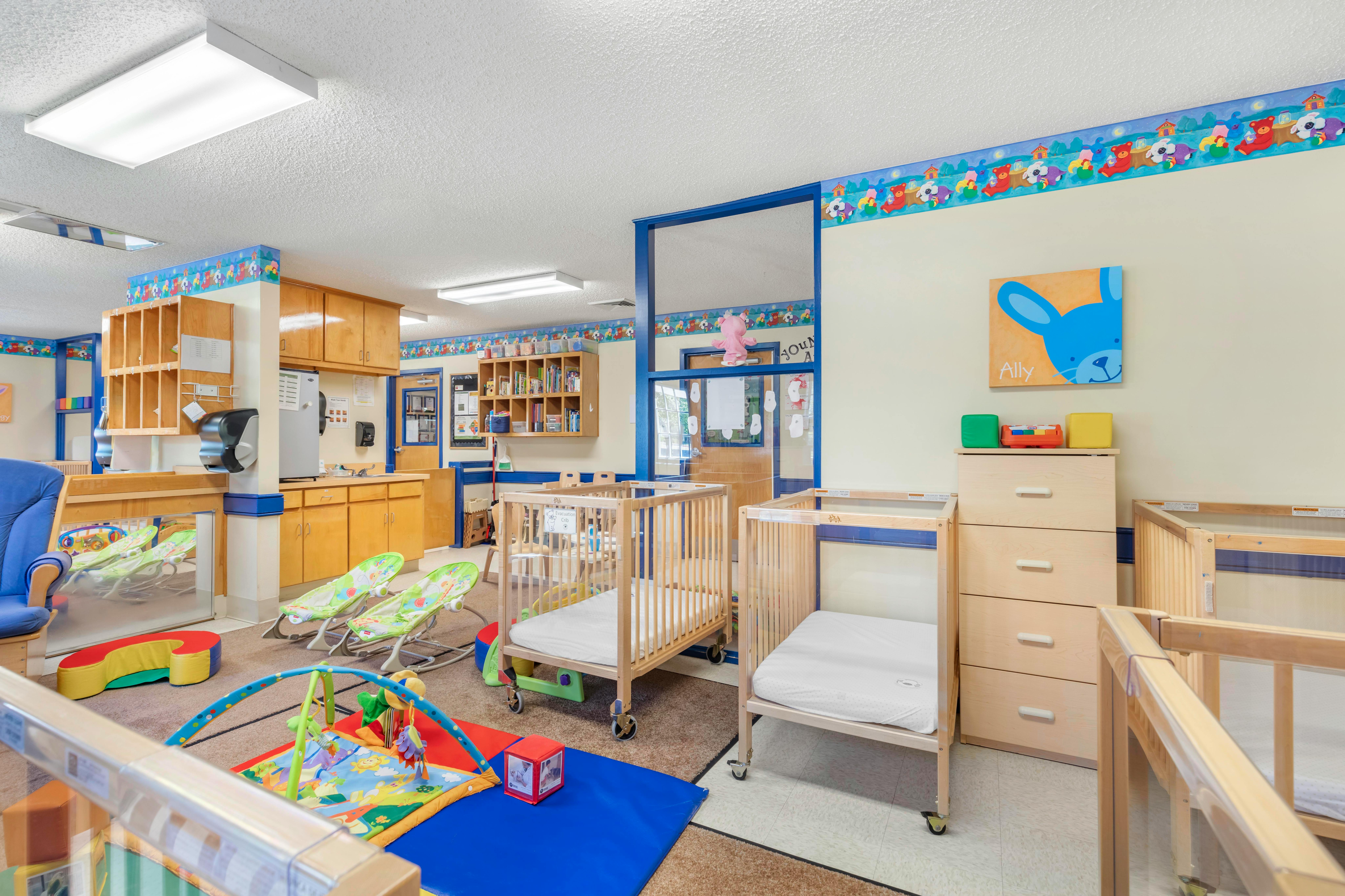 A colorful childcare classroom with several cribs, playmats, and baby bouncers