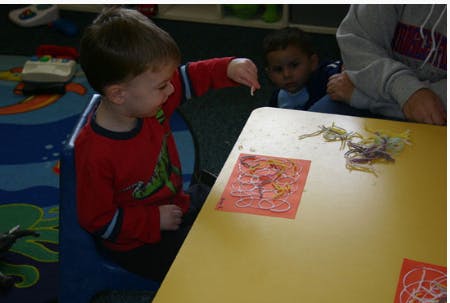 Child pointing at a paper on a table with another child watching