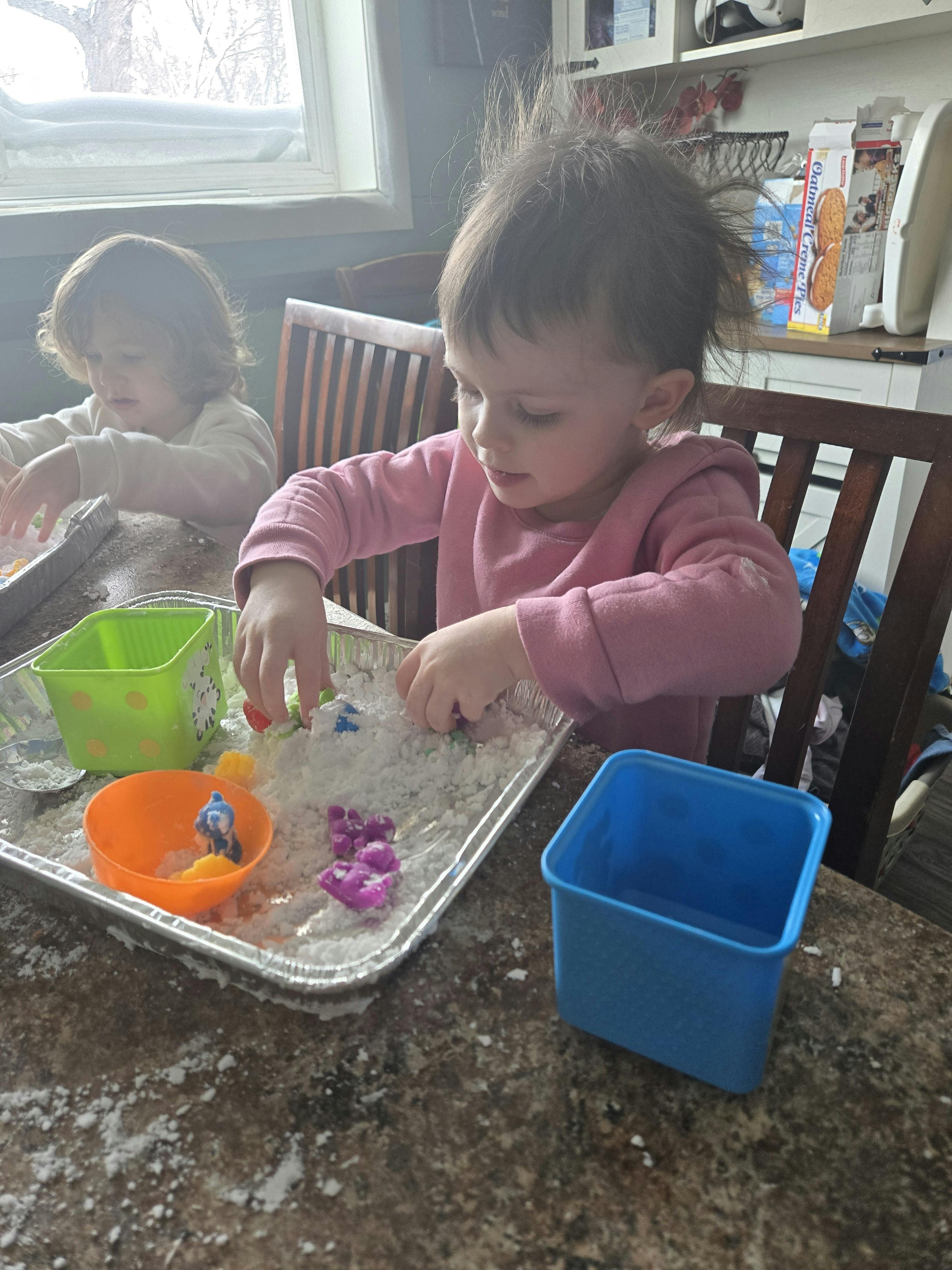 Two children playing with a tray of white material and colorful objects