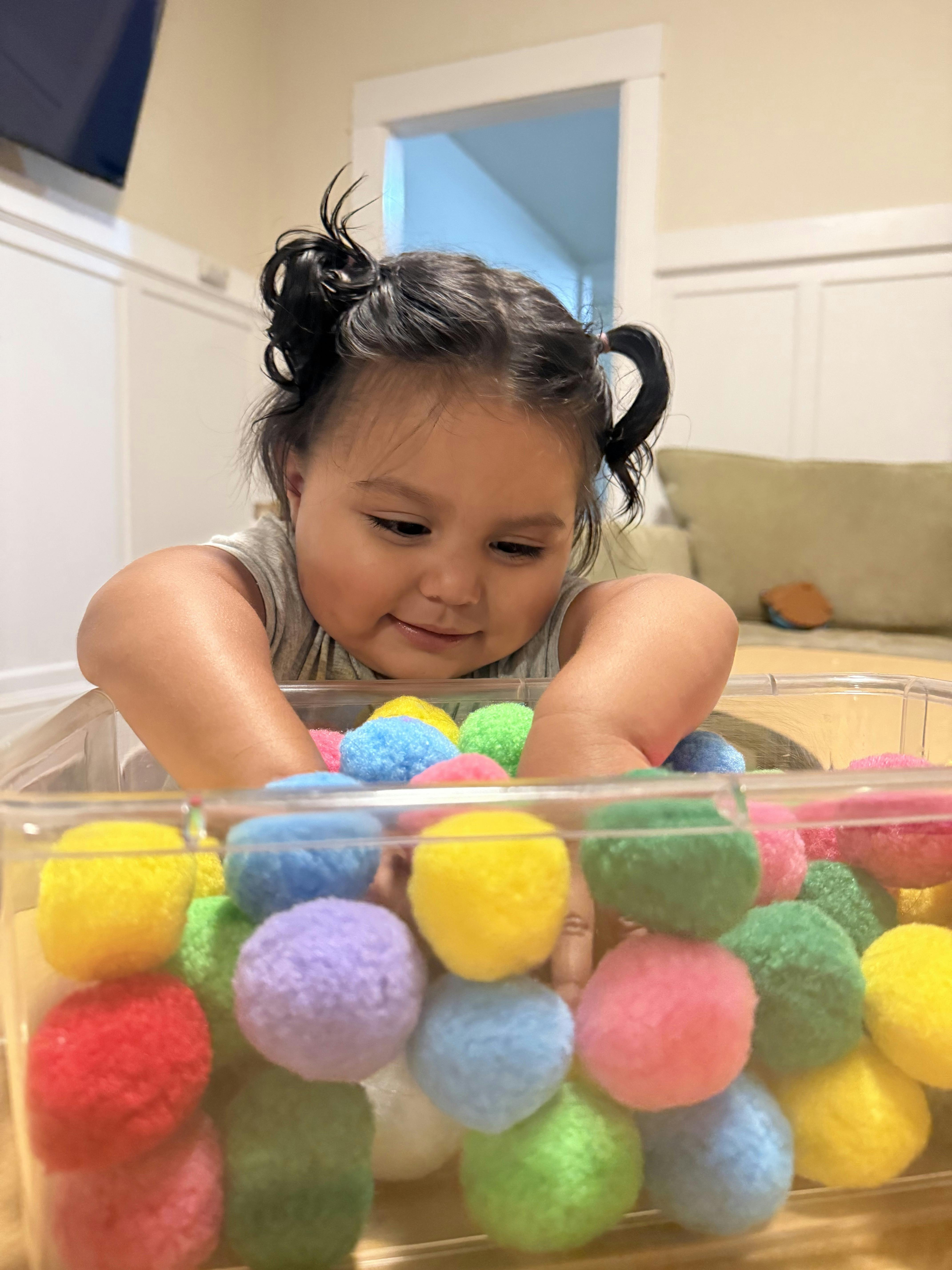 Child playing with colorful craft pom-poms in a clear plastic container