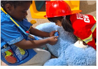 Two children playing with a teddy bear, one in a firefighter costume