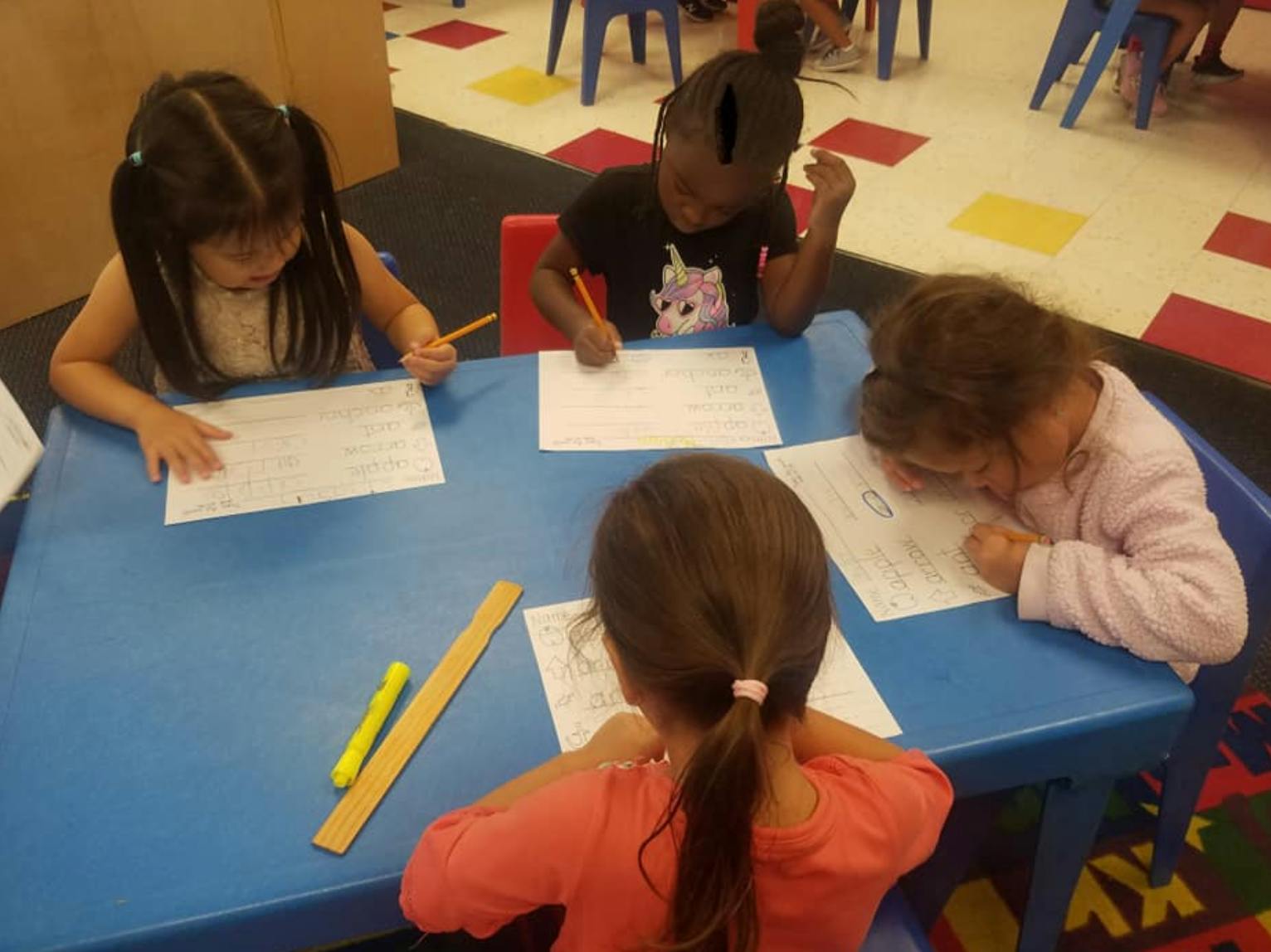 Four children writing at a blue table in a classroom