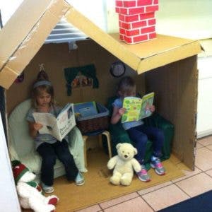 Two children reading books inside a cardboard playhouse