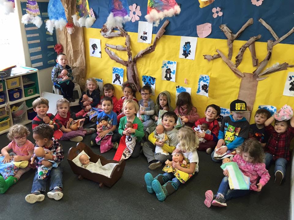 Many children sitting on the floor in a classroom holding dolls