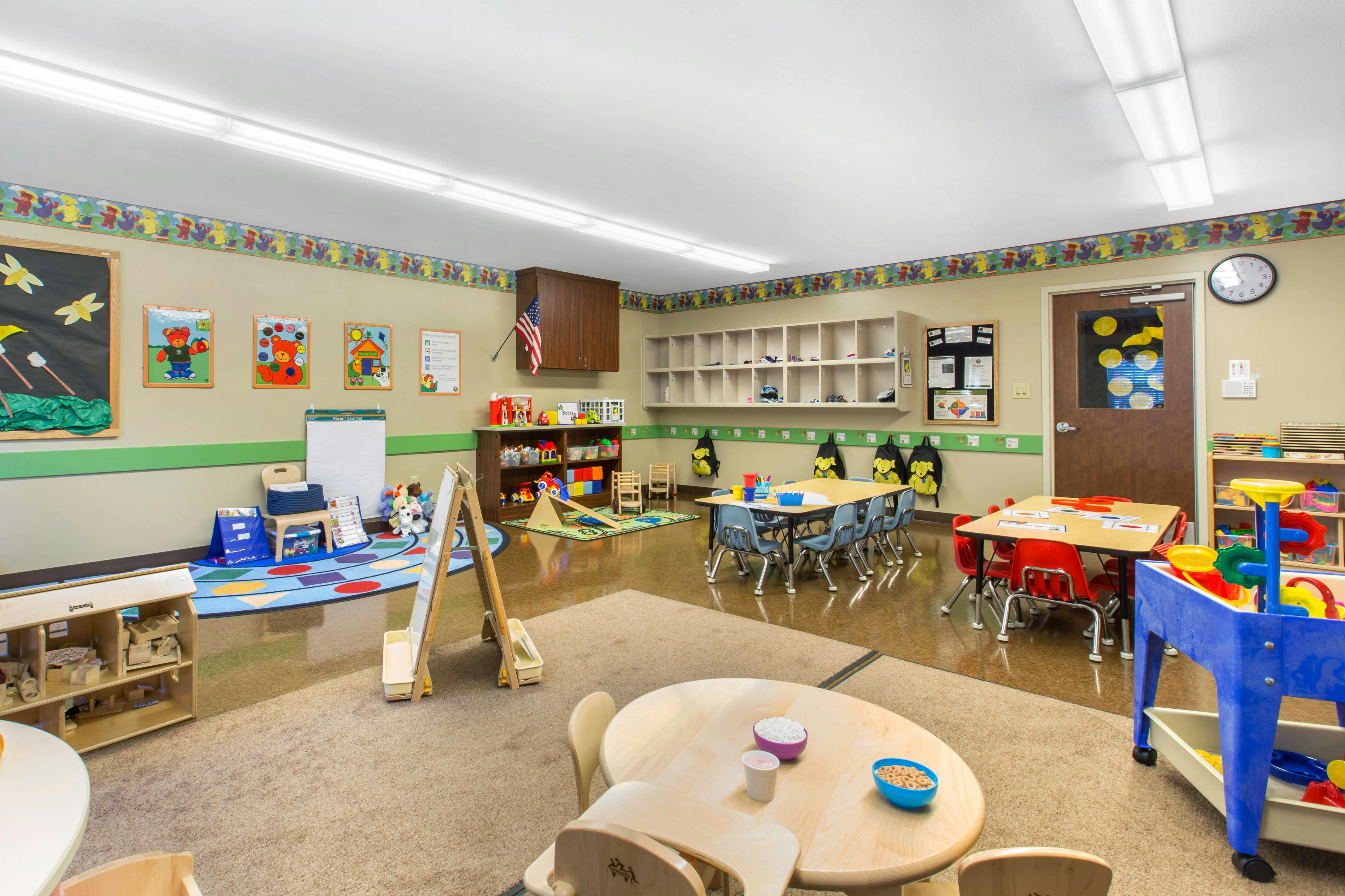 An empty classroom with small tables and chairs, play mats, and learning tools