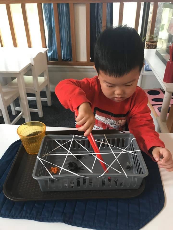 Child using a red tool to pick up objects from a basket with elastic bands