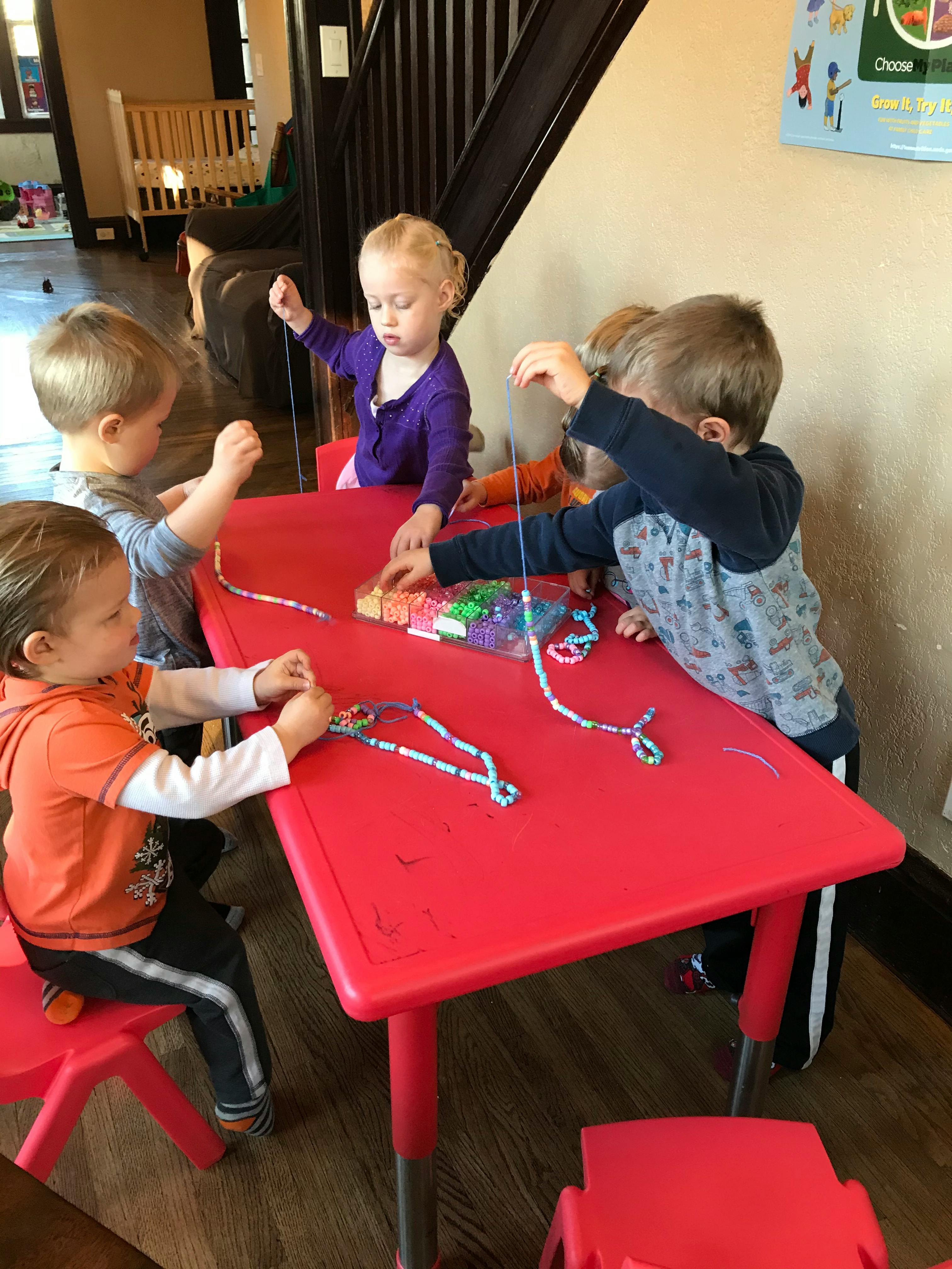 Children sitting at a red table stringing beads