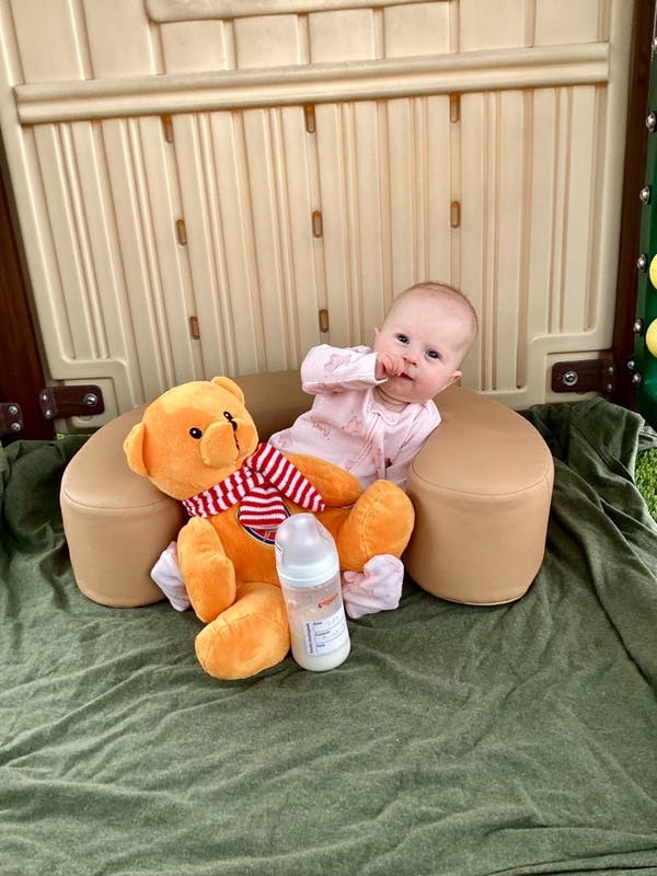 Baby sitting in a support cushion next to a teddy bear and a baby bottle