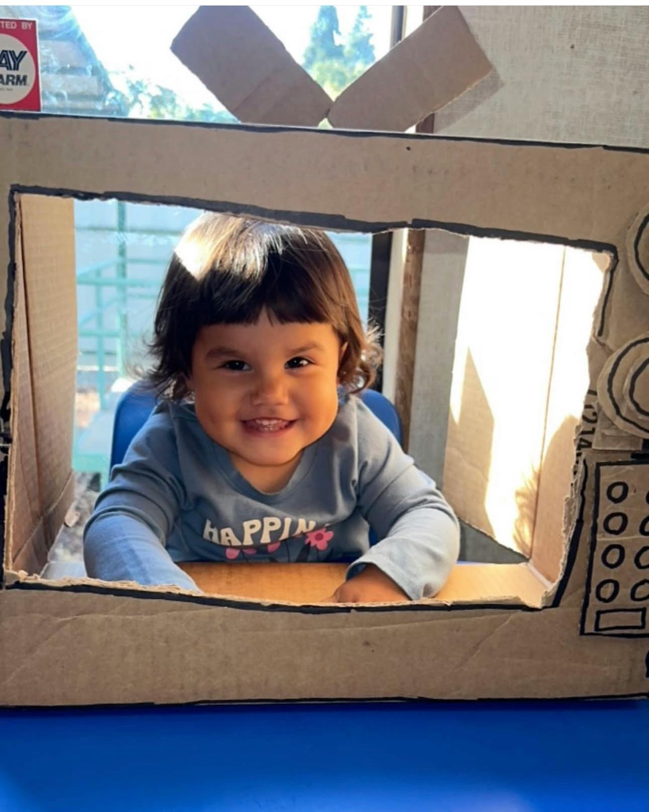 Child looking through a cardboard box frame made to look like a television