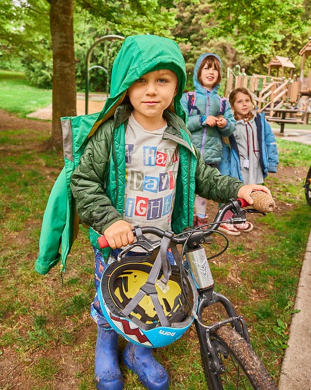 Child in a green jacket holding a bicycle with a helmet attached, two children behind