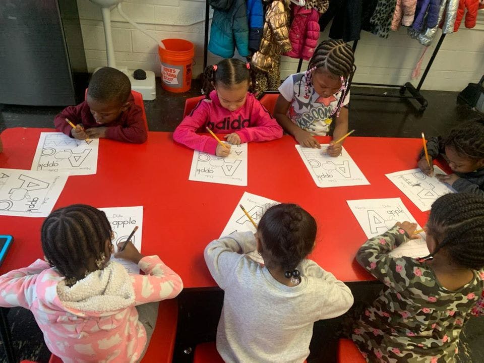 Children sitting at a red table, writing on papers with pencils on paper