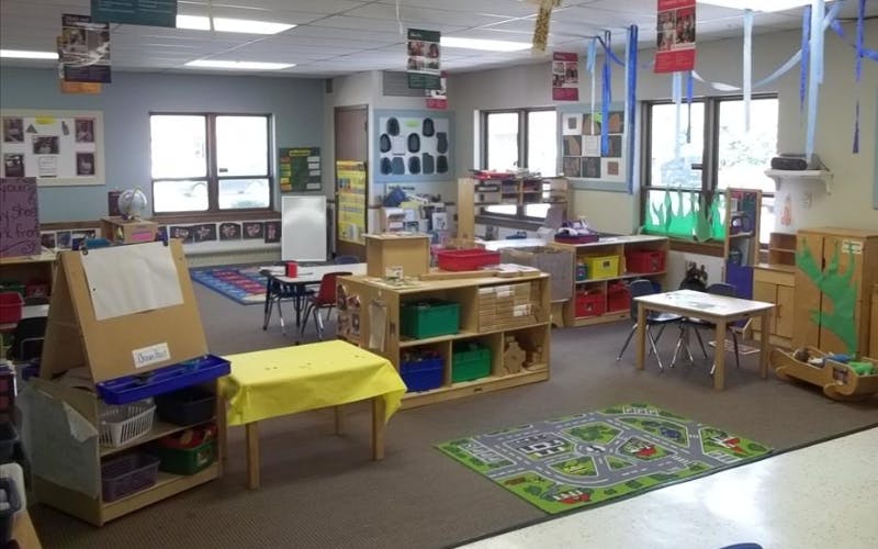 Classroom filled with small tables, chairs, shelving units with colorful bins, and rugs