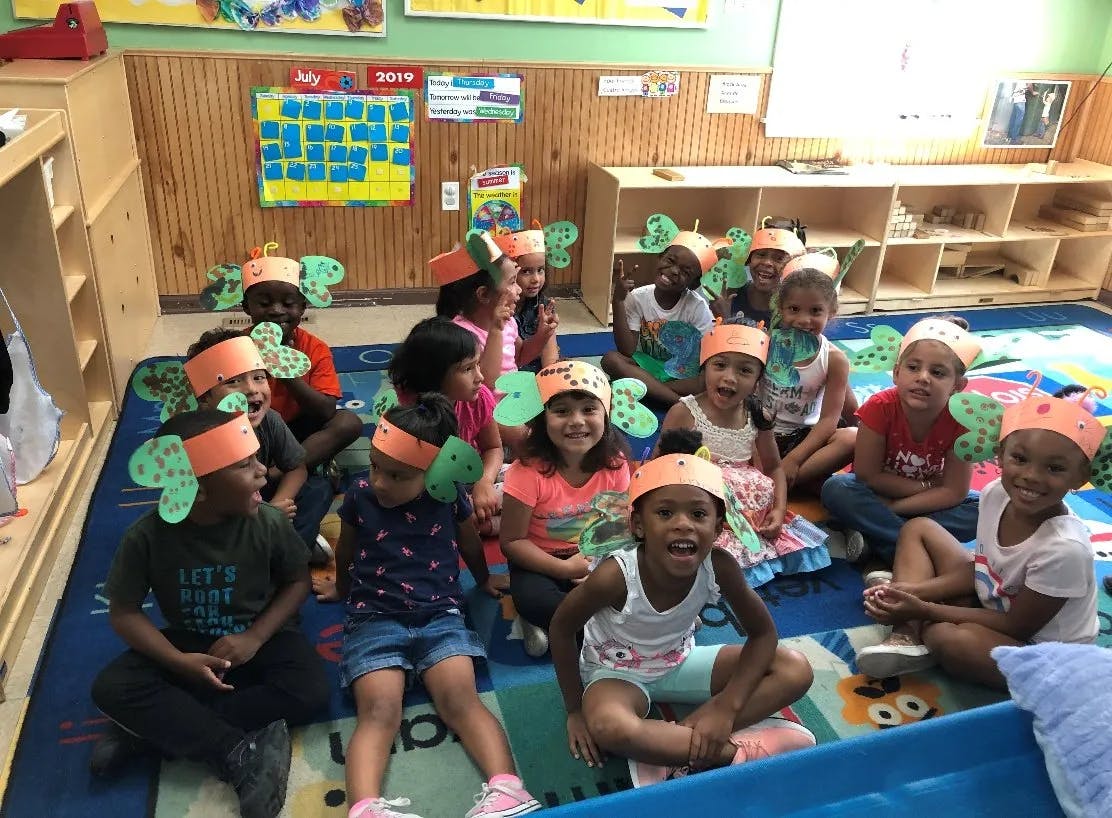 Group of children wearing paper hats with green cutouts sitting on a rug