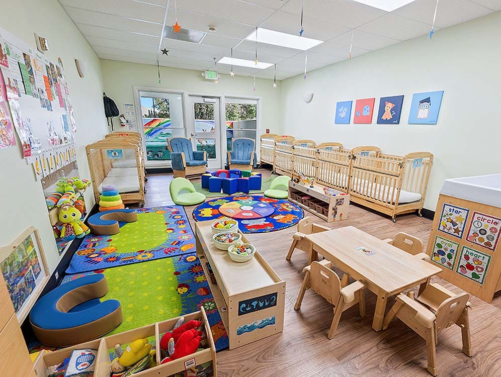 Colorful daycare or preschool classroom with cribs, play rugs, and child-sized furniture