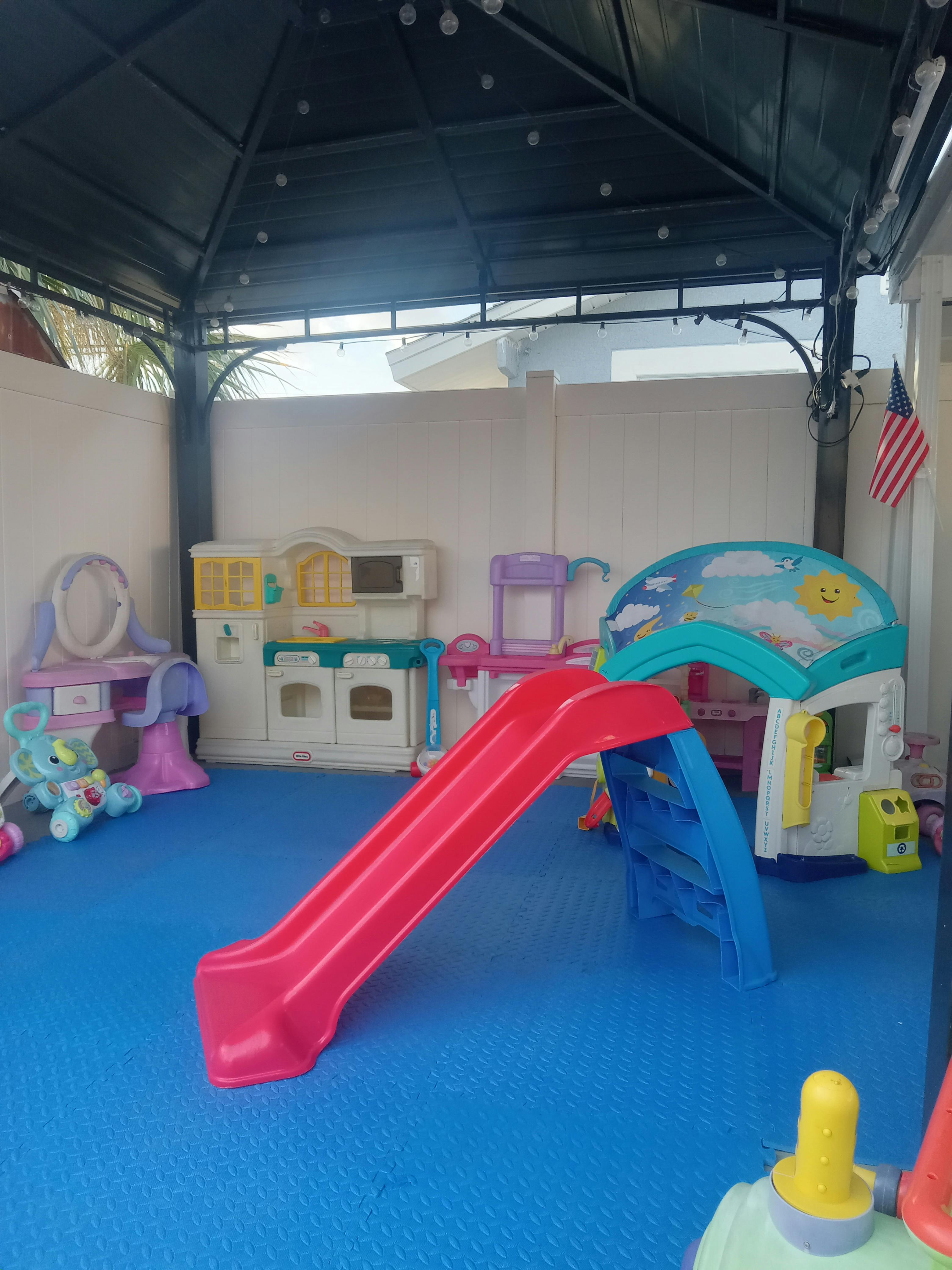 Playground with slide, play kitchen, and other toys on a blue mat under a gazebo