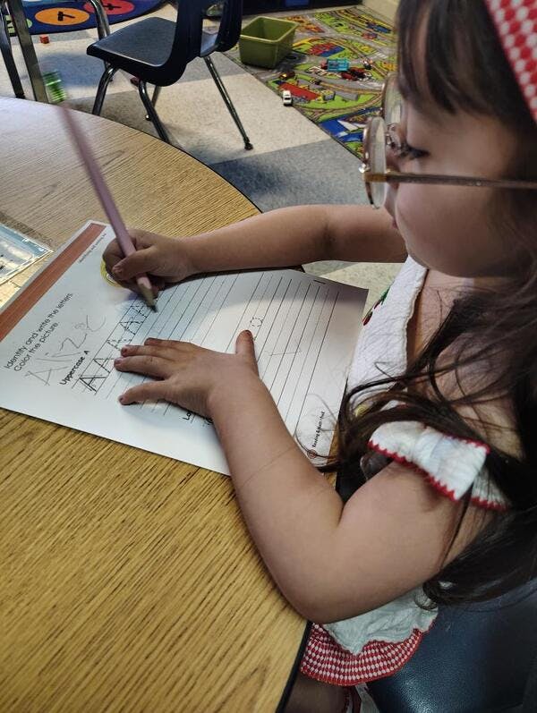 Child in glasses writing on a paper with a pencil at a wooden table