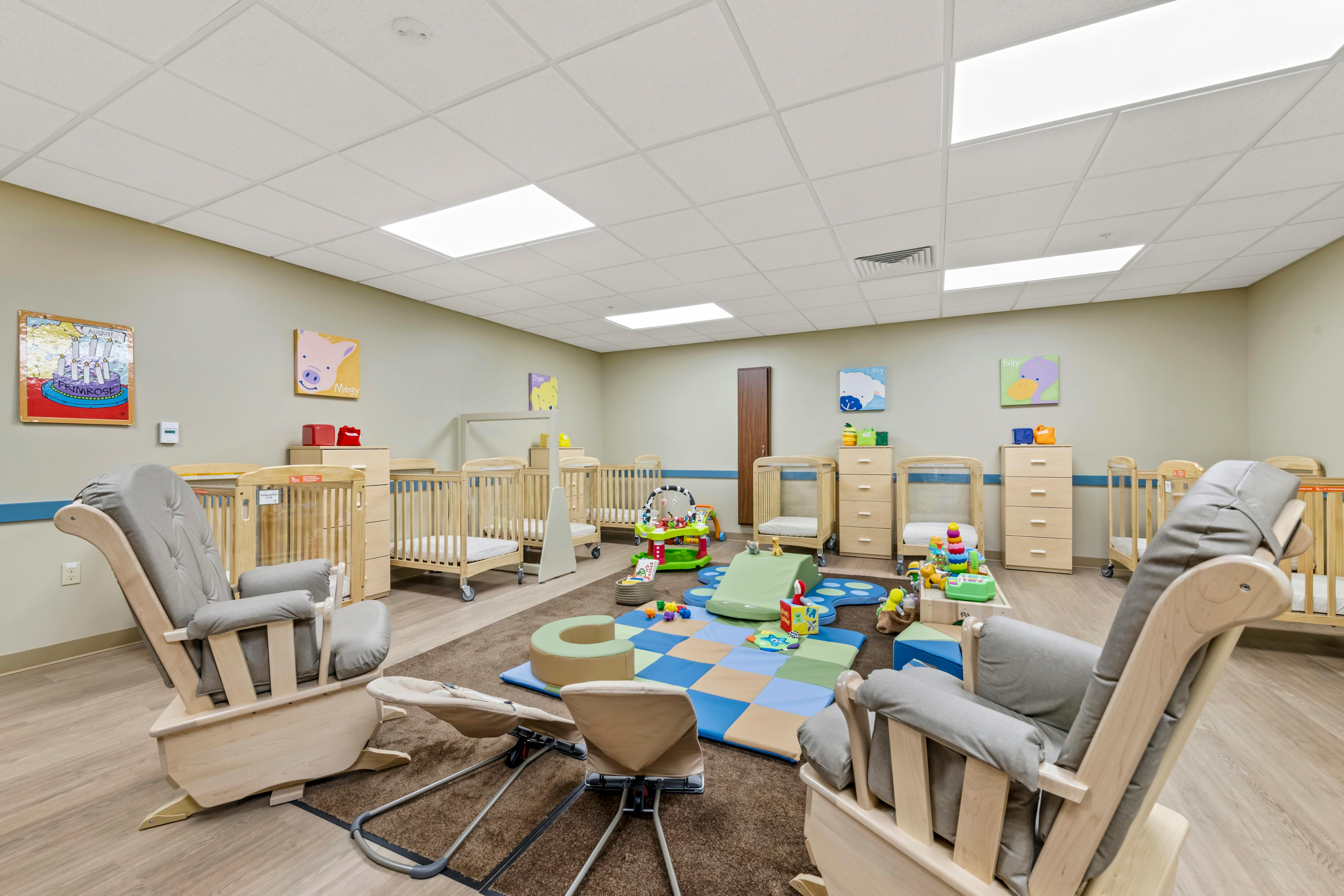 A childcare room with multiple cribs, a play area with soft mats, and two rocking chairs