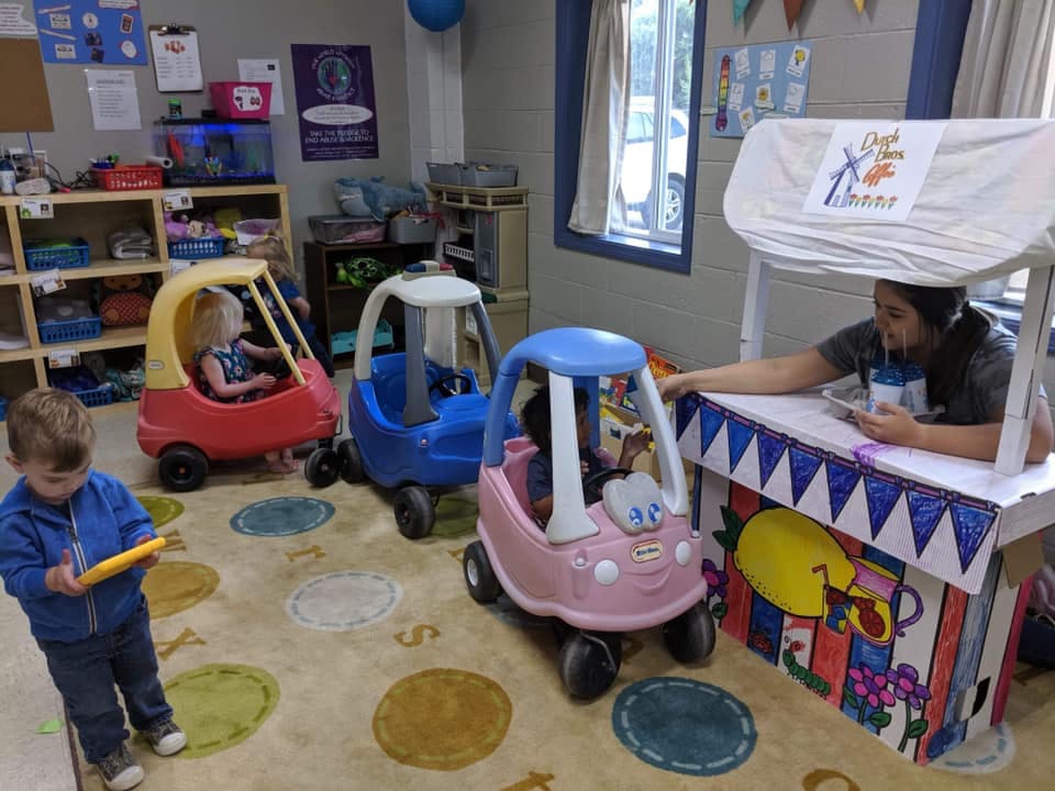 Children playing with toy cars and a person at a play stand in a classroom