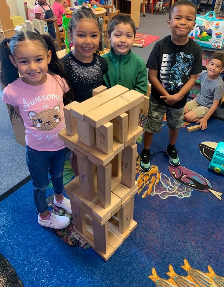 Four children standing around a tall structure made of wooden blocks
