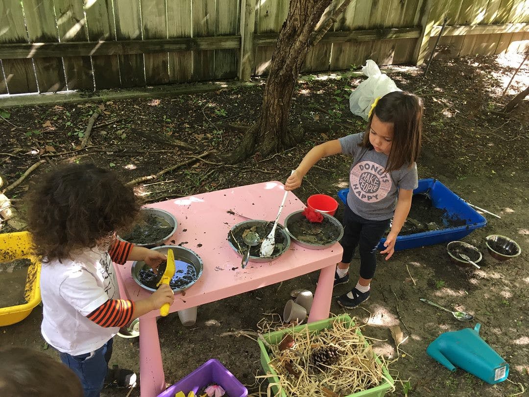 Two children playing with mud and bowls at a pink table outdoors