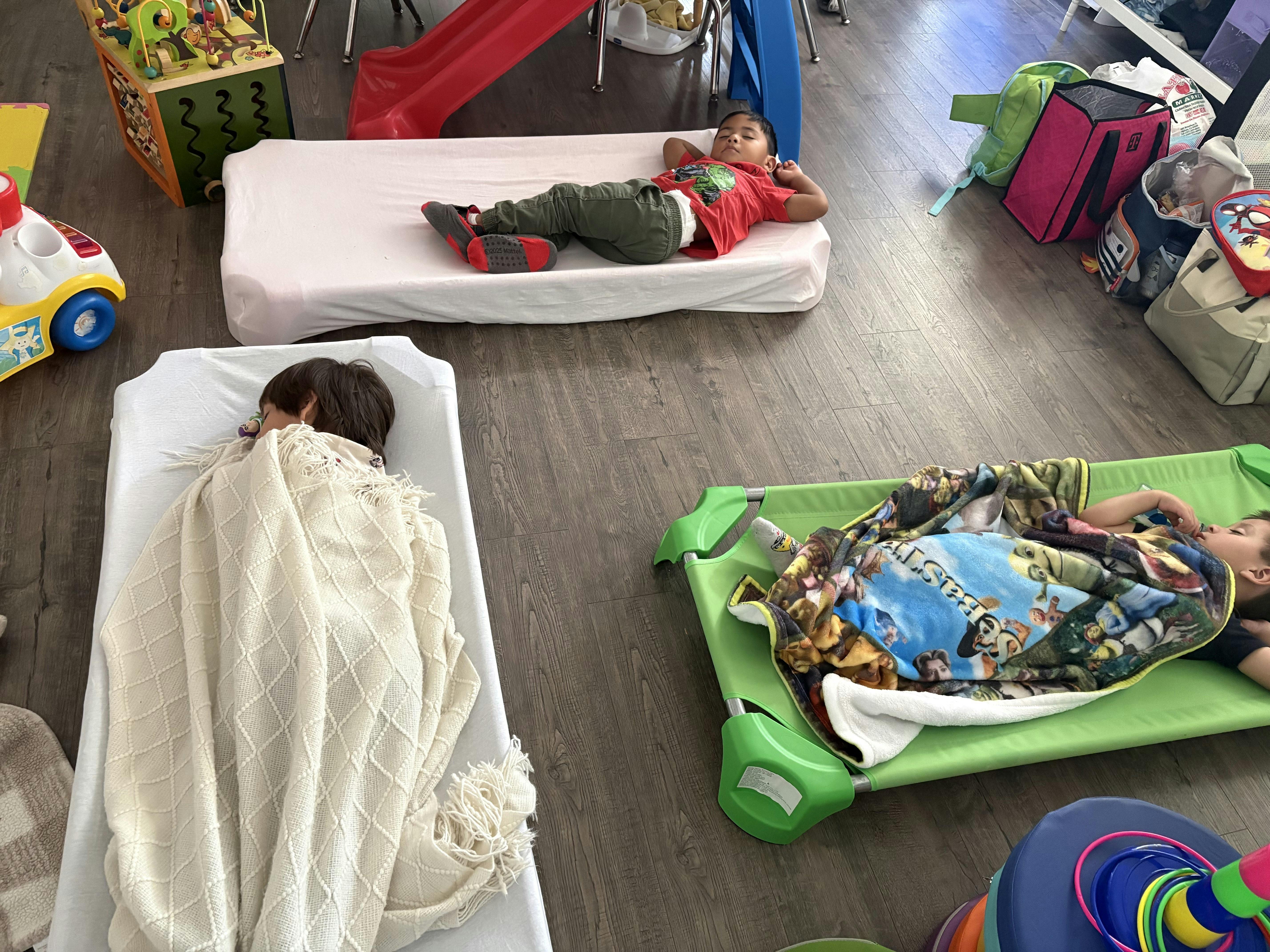 Three children sleeping on cots on a wooden floor in a classroom