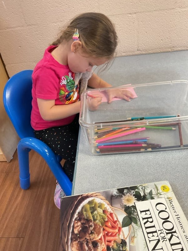 Child drawing on pink paper at a table with colored pencils