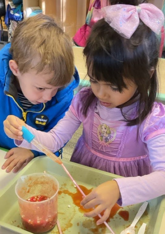 Two children using a dropper and straw with liquid in a tray