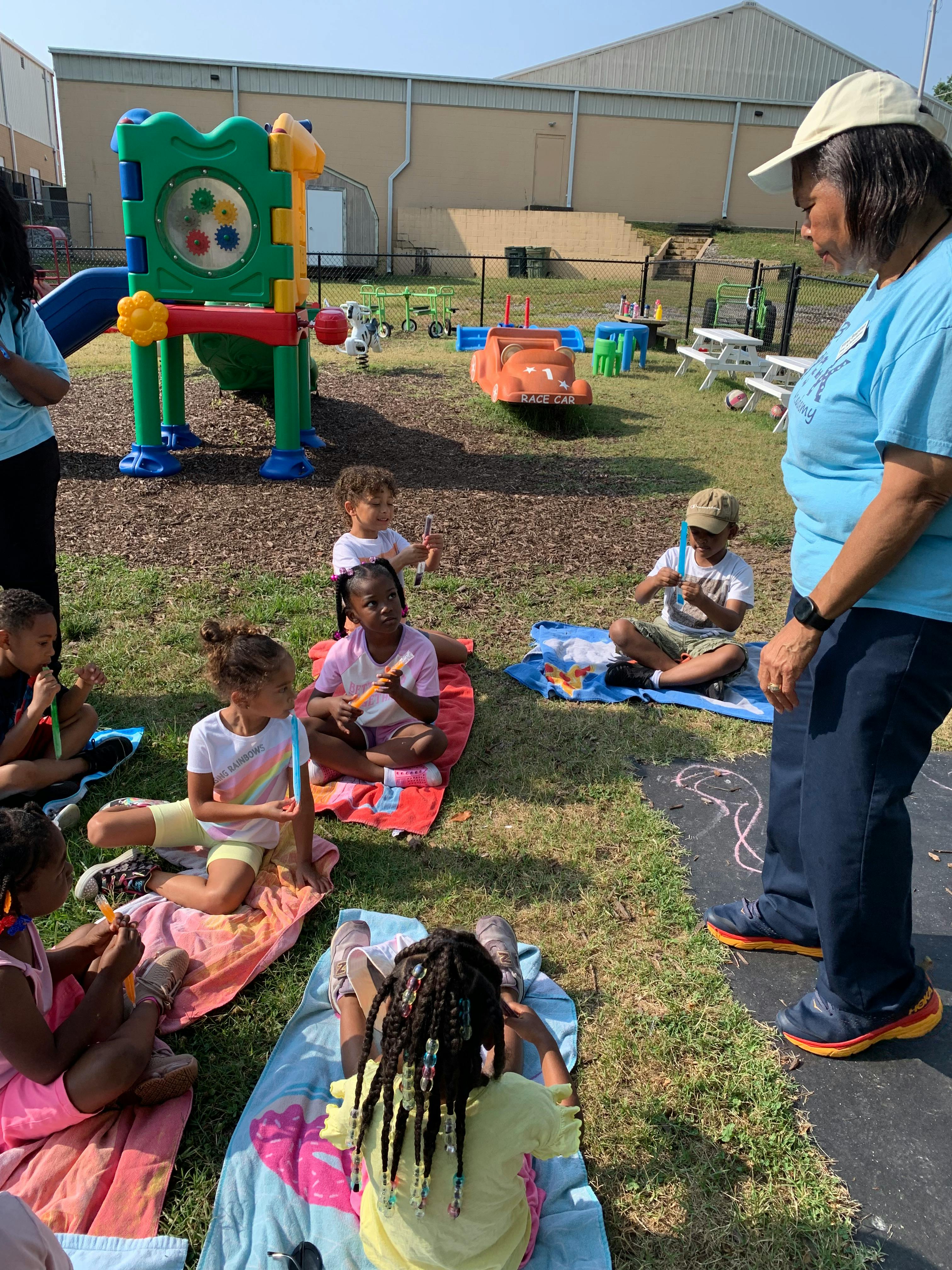 Adult standing near children sitting on blankets on grass in a playground