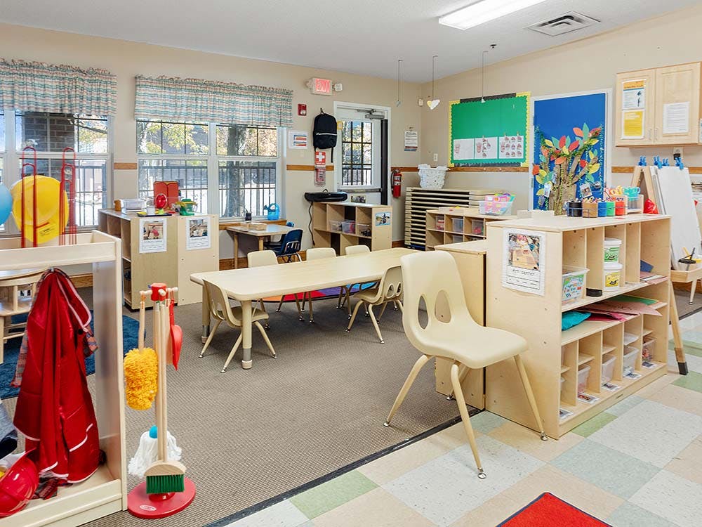 Empty classroom with small tables and chairs, shelves with supplies, and windows